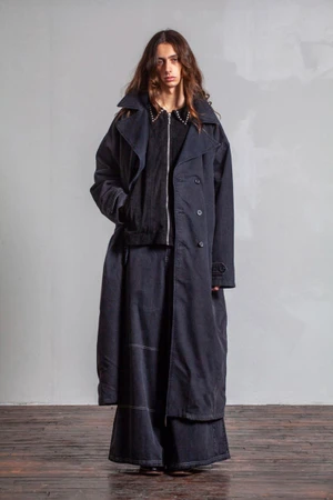 Model with long wavy hair wearing a dark trench coat over a zipped jacket and wide trousers against a grey wall