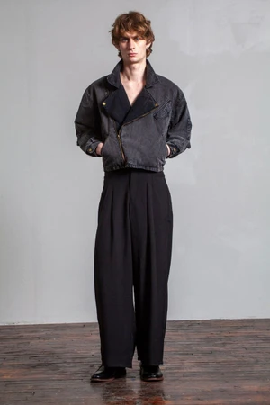 Full-length portrait of a male model wearing a dark denim jacket and pleated black trousers against a neutral wall
