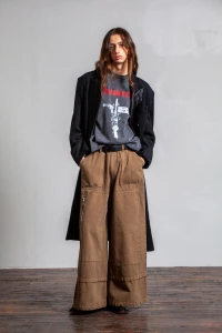 Male model with long hair wearing a black coat and brown cargo pants against a plain wall and wooden floor