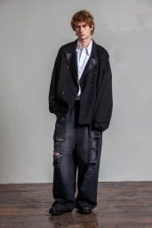 Model in oversized black double-breasted blazer and distressed dark jeans standing against a grey wall.