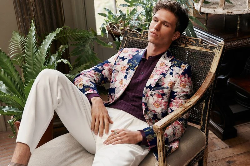 Man in a floral blazer sits in a rattan chair with a relaxed pose. Indoor setting with houseplants and soft, diffused lighting.