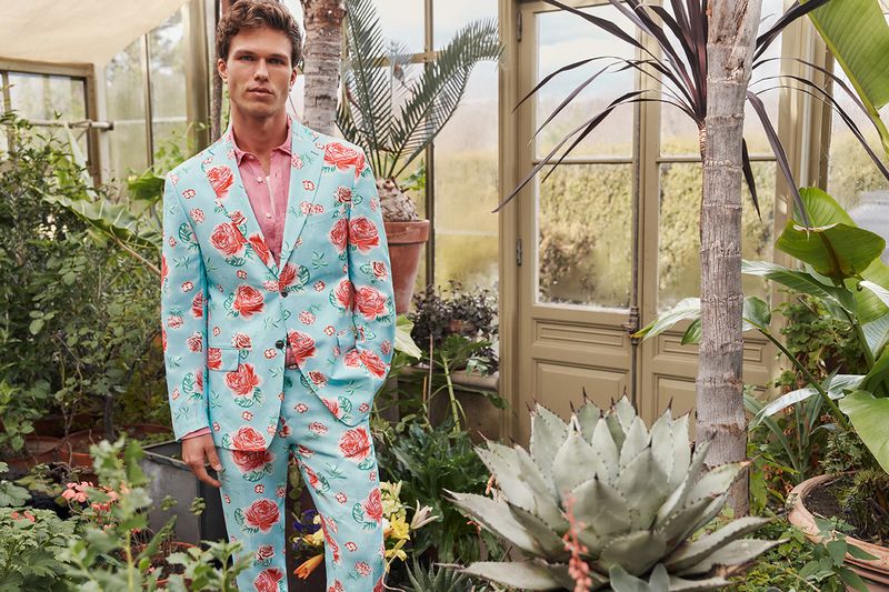 Male model in a rose-patterned suit poses inside a glasshouse filled with lush greenery and plants, creating a vibrant, fashionable scene.