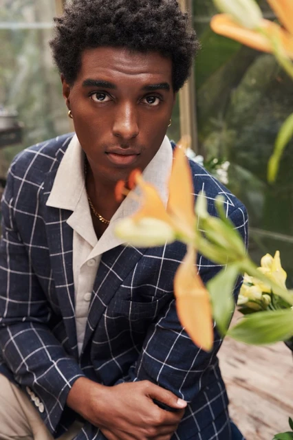 Close-up portrait of a Black man with dark hair wearing a navy plaid blazer with orange flowers in foreground, looking directly at the camera.