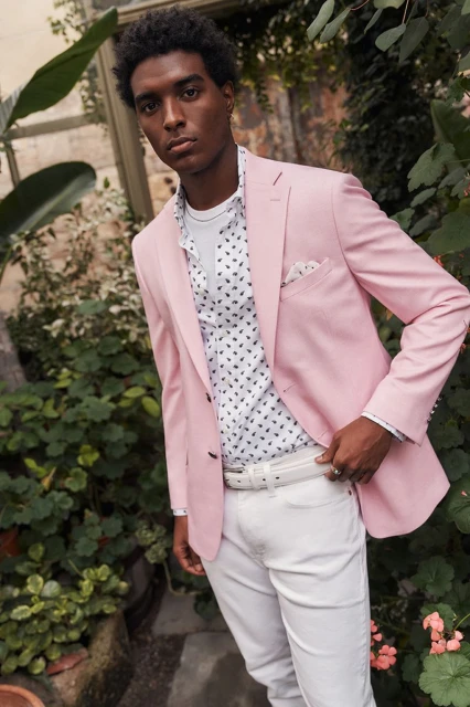 A young man wearing a pink blazer and white outfit stands in a botanical garden.