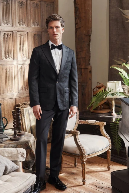 Elegant man in a black suit with a bow tie standing indoors in front of a decorative wooden wall and chair with leafy plants.