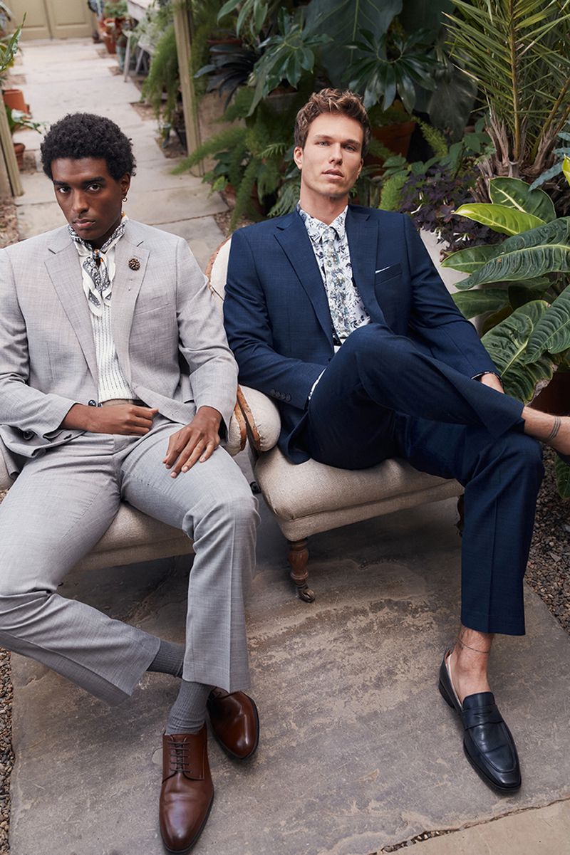 Two adult men pose indoors wearing tailored suits and dress shoes, seated on vintage chairs amongst potted green plants.
