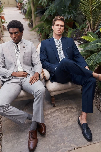 Two adult men pose indoors wearing tailored suits and dress shoes, seated on vintage chairs amongst potted green plants.