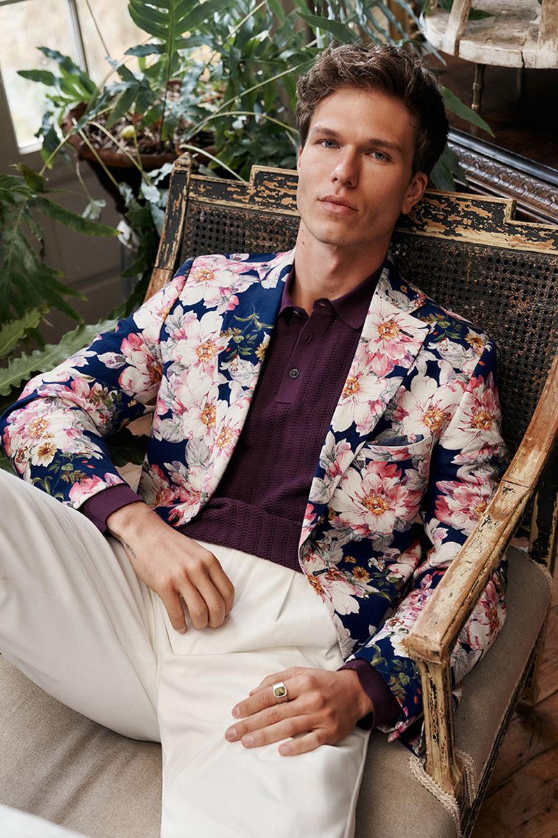 Man wearing a navy blue floral blazer and purple polo shirt sits in an antique cane chair, looking at the camera.