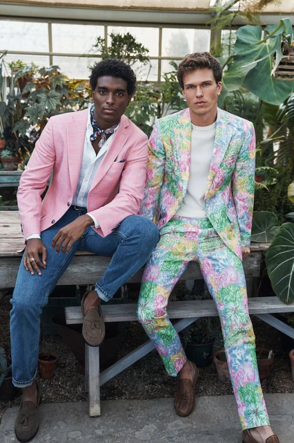 Two stylish men pose outdoors in floral and pink blazers with jeans and loafers, surrounded by lush greenery in a greenhouse setting.