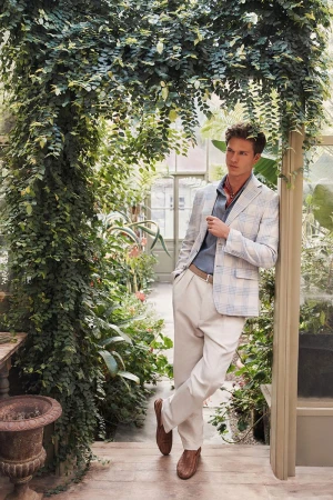 A man wearing a light plaid blazer and cream trousers leaning against a greenhouse pillar framed by lush vines.