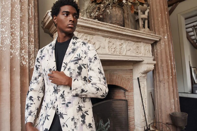 Man in floral suit jacket poses indoors near fireplace. He wears a black t-shirt and faces to the left, with columns visible on left.