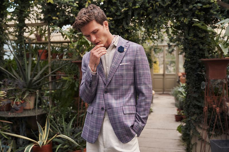 Man poses in a plaid blazer and cream-colored pants inside a sun-drenched greenhouse filled with potted plants and natural light.