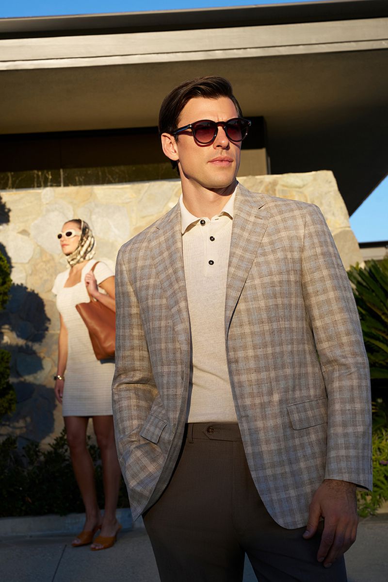Male model in plaid jacket poses with a woman in a dress and headscarf near a modern building on a sunny day, fashion photography.