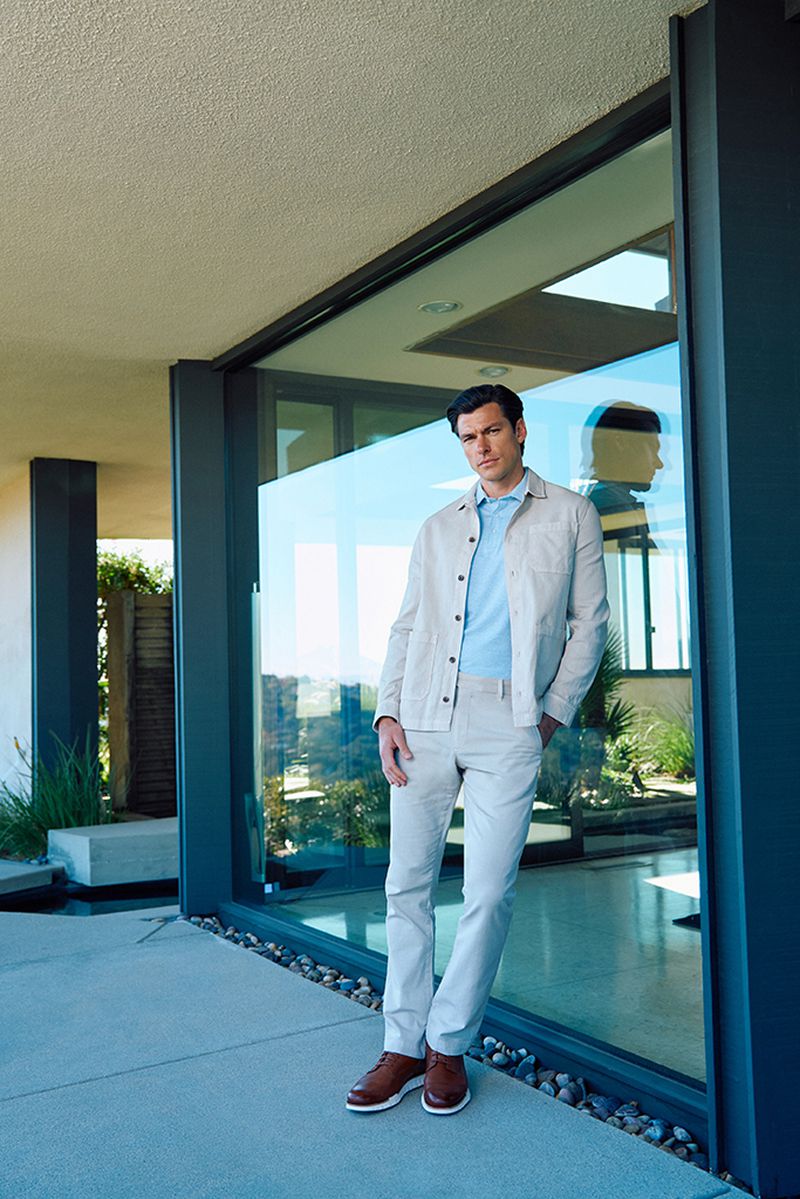 Man in a light-colored casual outfit standing by a glass wall outdoors, looking at the camera with neutral expression. Light and airy.