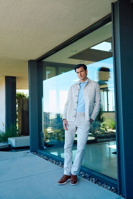Man in a light-colored casual outfit standing by a glass wall outdoors, looking at the camera with neutral expression. Light and airy.