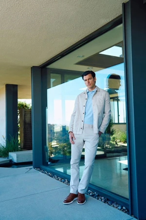 Man in a light-colored casual outfit standing by a glass wall outdoors, looking at the camera with neutral expression. Light and airy.