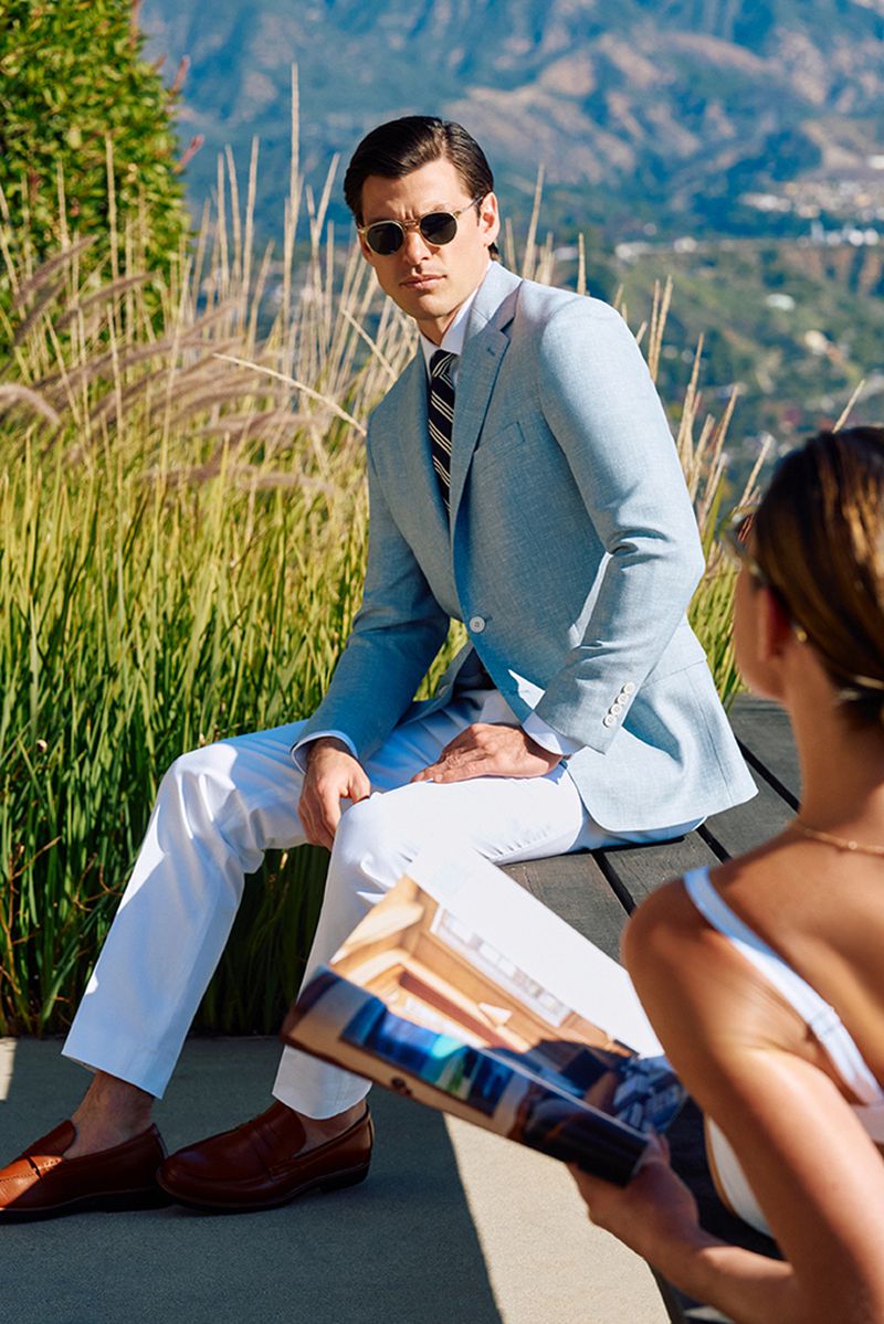 A man in a light blue suit sits on a bench with a woman reading a magazine on a sunny day in a landscaped setting with a mountain backdrop.