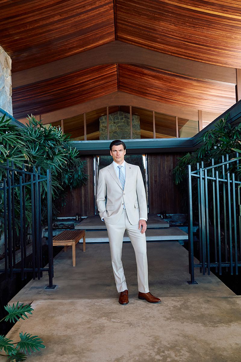 An adult male stands in a beige suit outside a modern, wood-clad building surrounded by greenery and wrought iron fencing, portrait shot.
