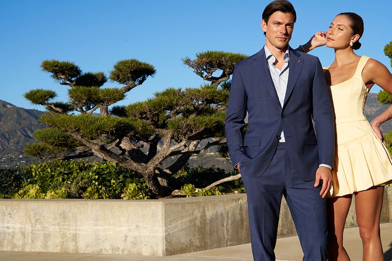 A man and woman pose outdoors, the man wears a blue suit, and the woman wears a yellow dress; mountains and greenery are in the background.