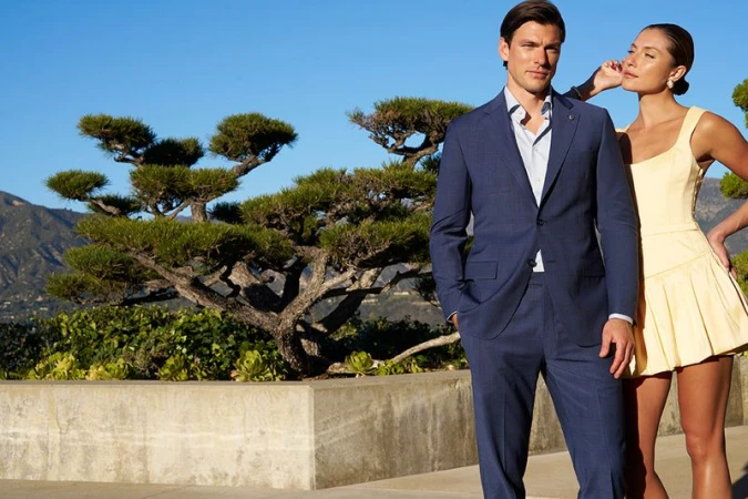 A man and woman pose outdoors, the man wears a blue suit, and the woman wears a yellow dress; mountains and greenery are in the background.