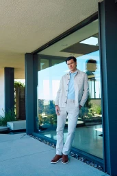 Man wearing a light beige jacket and matching trousers leaning against a glass wall of a modern building