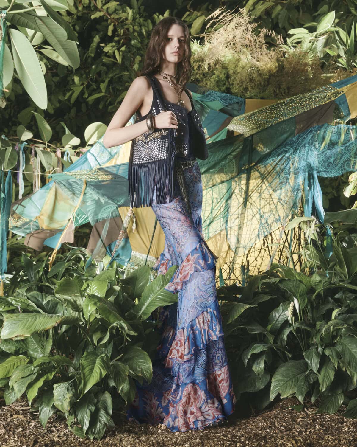 A woman in a printed sleeveless top and full-length skirt stands outdoors surrounded by greenery and hanging patterned textiles, bohemian fashion.