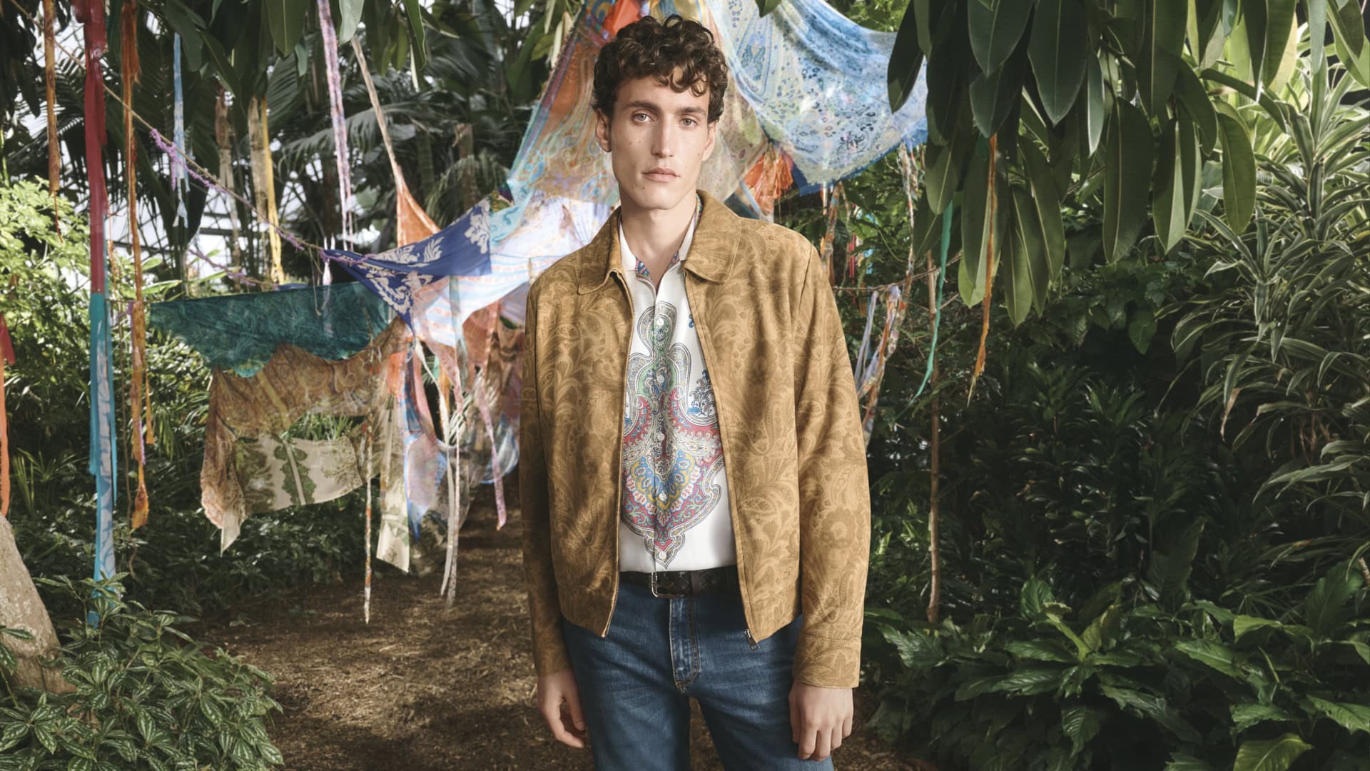 An adult male stands in a paisley jacket and jeans, surrounded by a lush botanical garden with draped fabrics overhead.