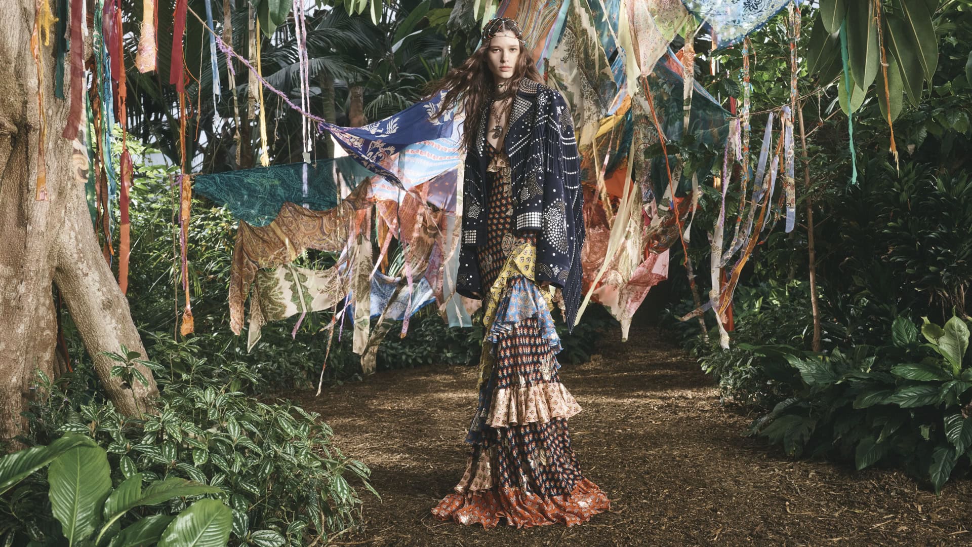 A fashionable woman in a long, tiered dress and jacket stands in a lush forest setting, with colorful textile decorations.