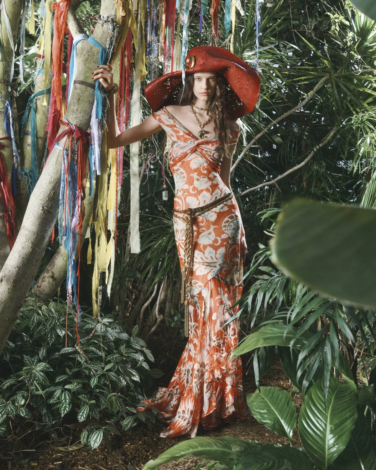 A stylish woman in an orange floral dress and wide-brimmed hat poses amidst lush tropical foliage and colorful ribbons tied to a tree.