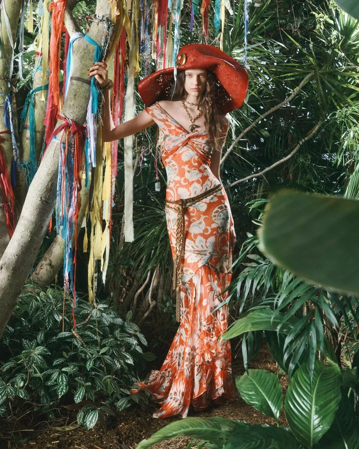 Model in orange patterned dress and wide red hat leaning against a tree decorated with colorful ribbons