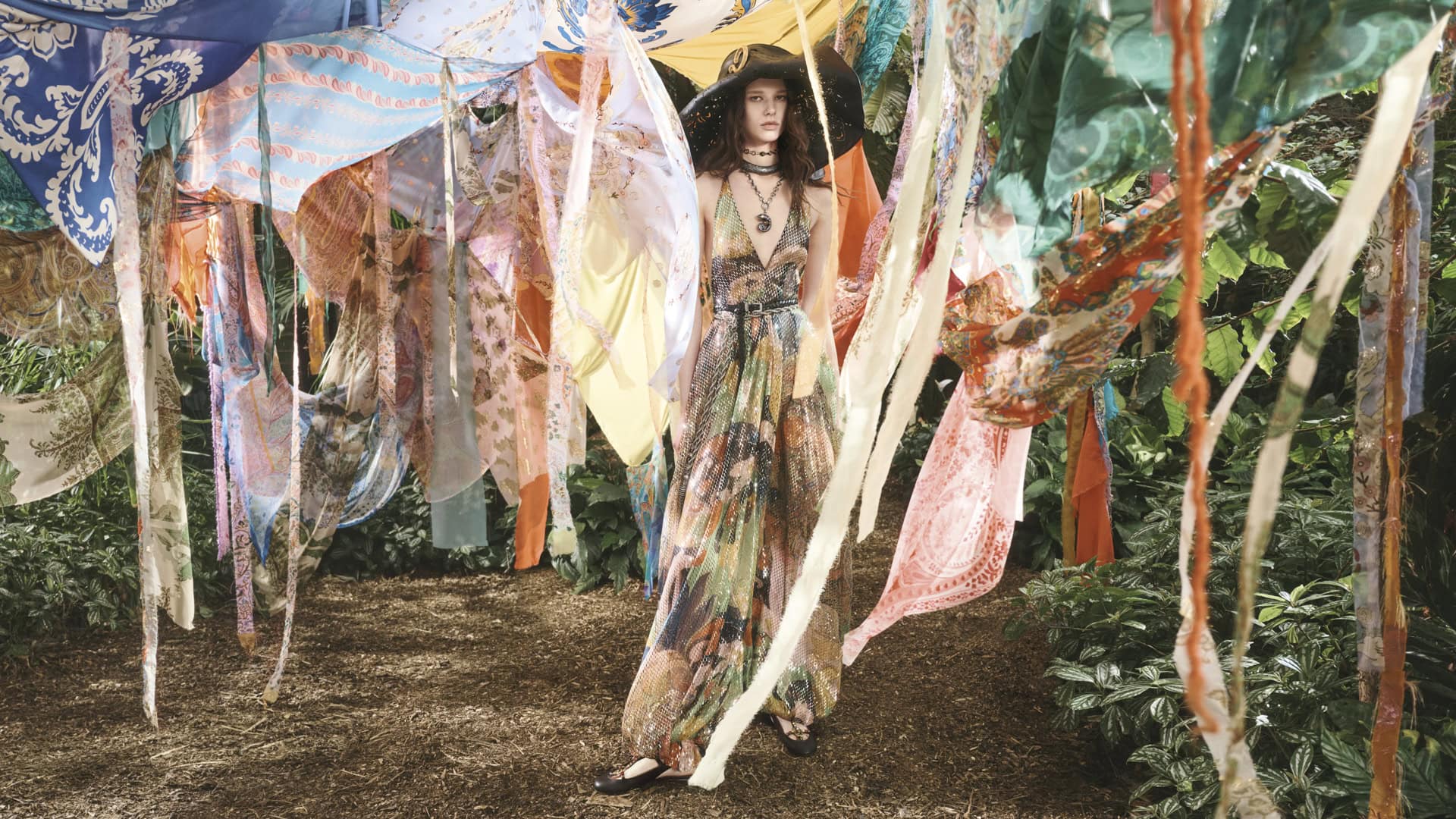 Model wearing patterned halter dress and wide-brimmed hat amid hanging colorful fabrics and lush foliage