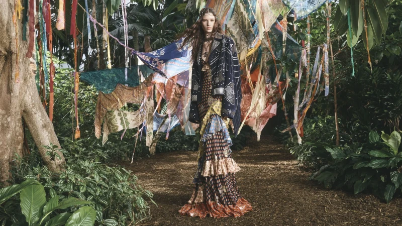 Model standing on a dirt path in a lush garden surrounded by hanging patterned textiles for Etro