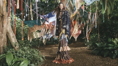 Model standing on a dirt path in a lush garden surrounded by hanging patterned textiles for Etro