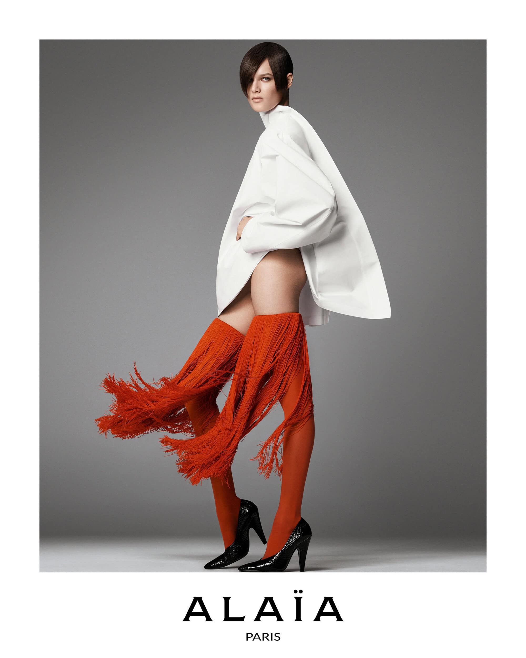 Fashion portrait of a woman with short brown hair wearing an oversized white jacket, red fringe socks, and black heels on a grey background.