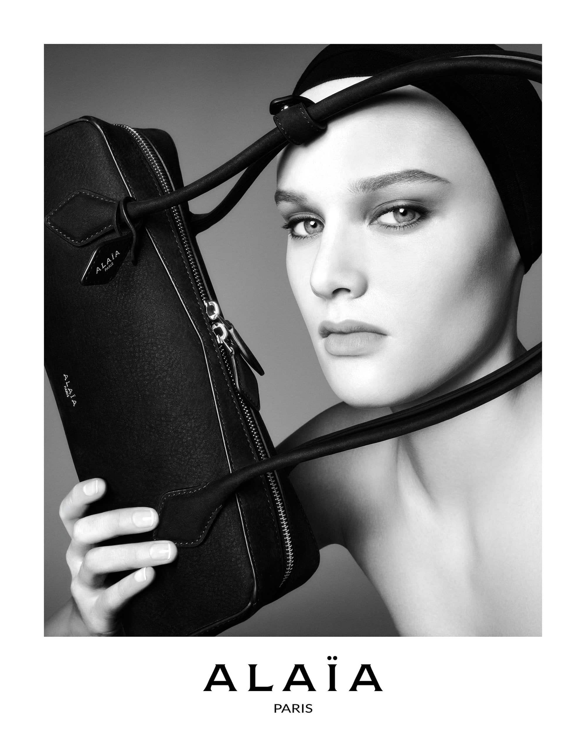 Black and white studio shot of a model holding a black leather Alaïa handbag with a black head covering and matching shoulder strap.
