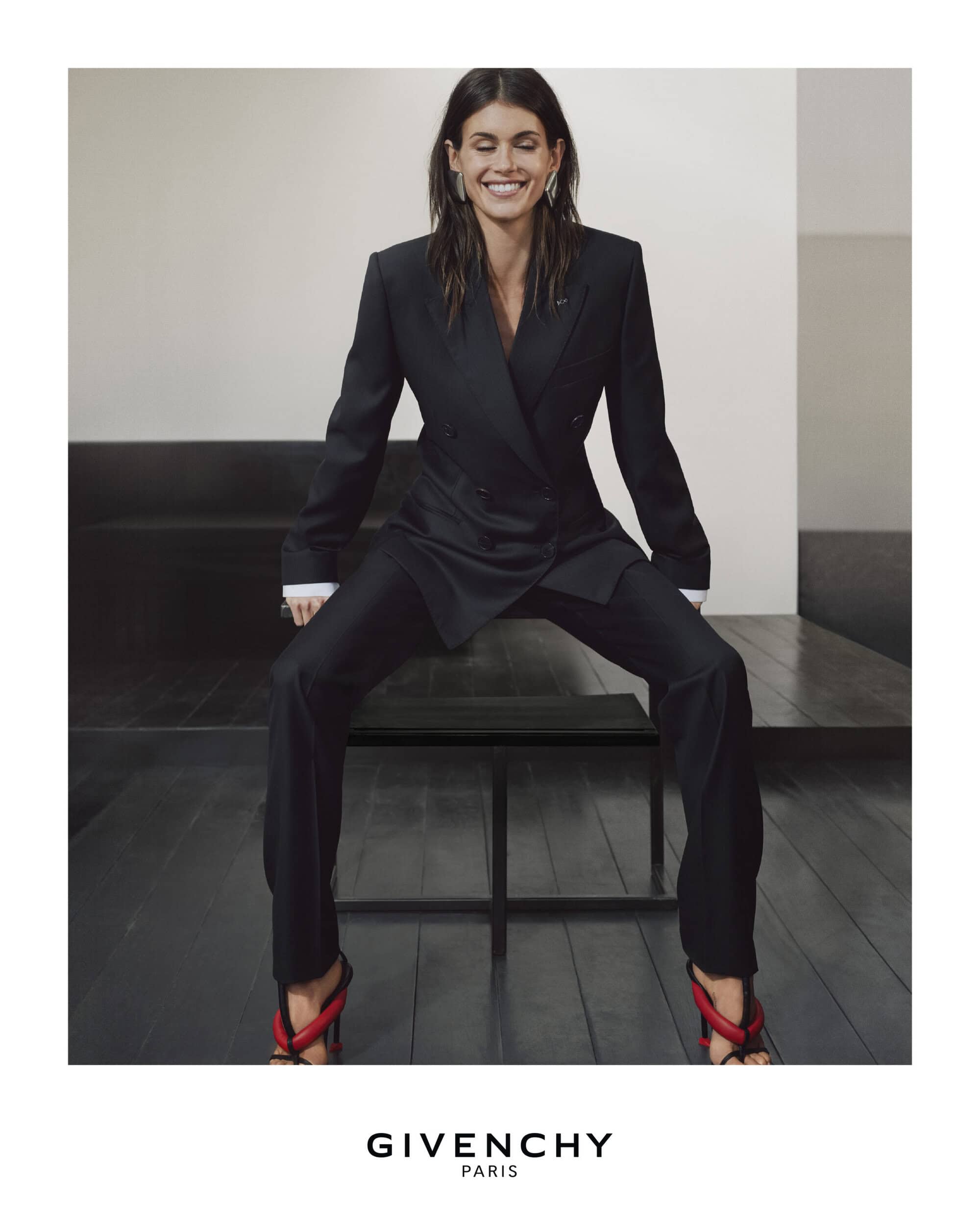 A smiling woman with dark hair poses in a dark double-breasted suit and red heeled sandals, sitting on a black bench against a white wall.