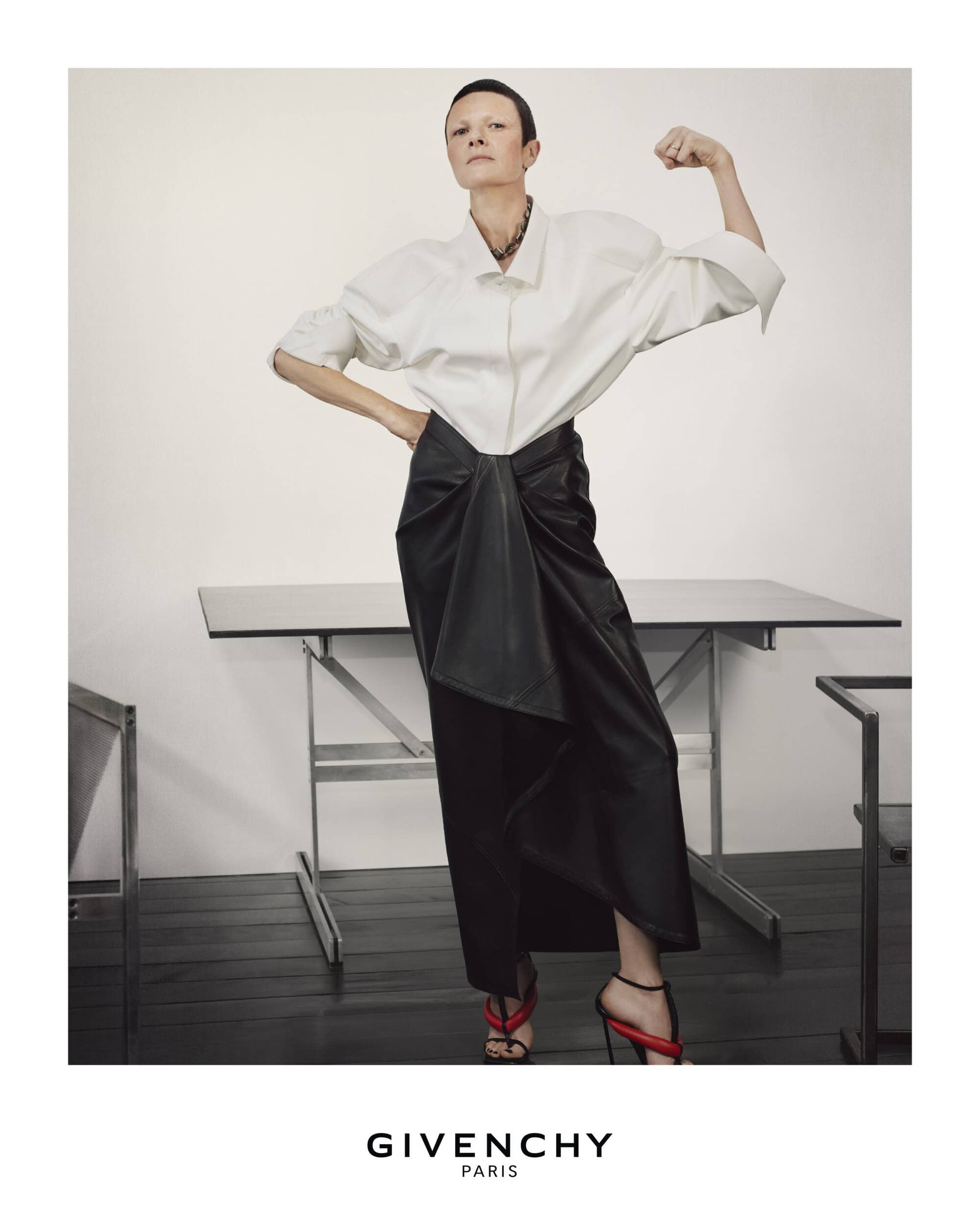Fashion shot of an adult woman with short hair, wearing a white shirt and black skirt with heels, flexing her arm in a studio.