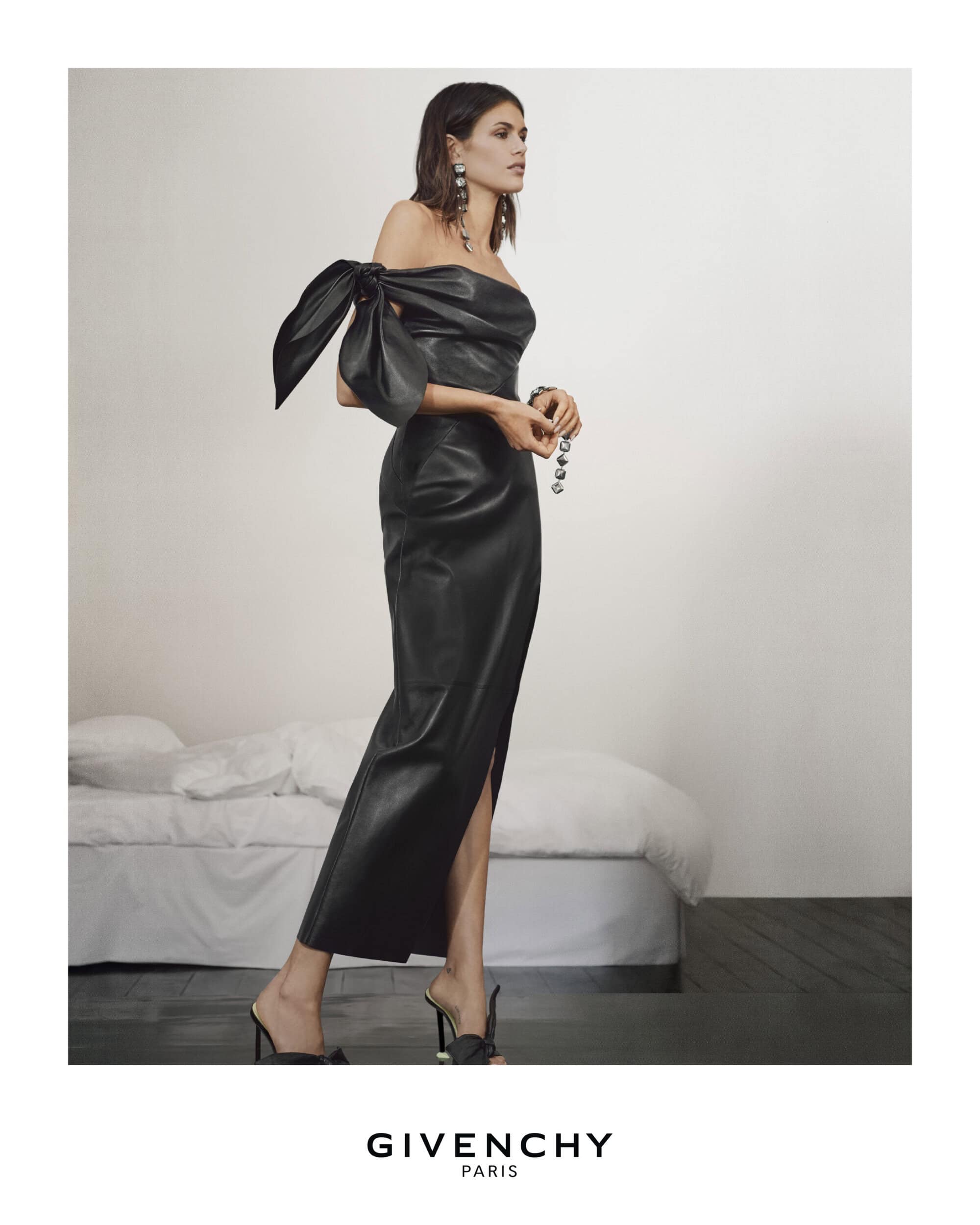 A woman in a black leather dress and heels poses in a minimalist studio setting, showcasing high-fashion apparel with elegant accessories.