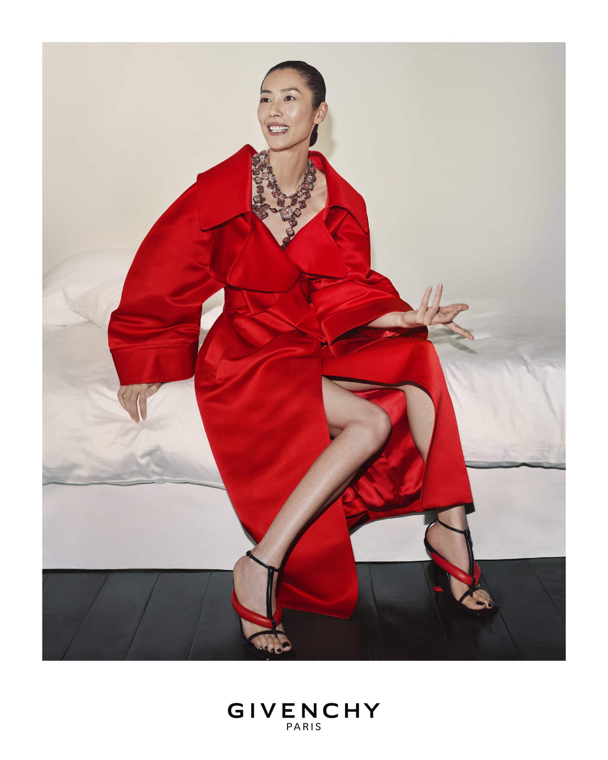 Elegant woman poses in a red dress and jewelry while sitting on a bed in an indoor setting. Fashion and beauty advertising.