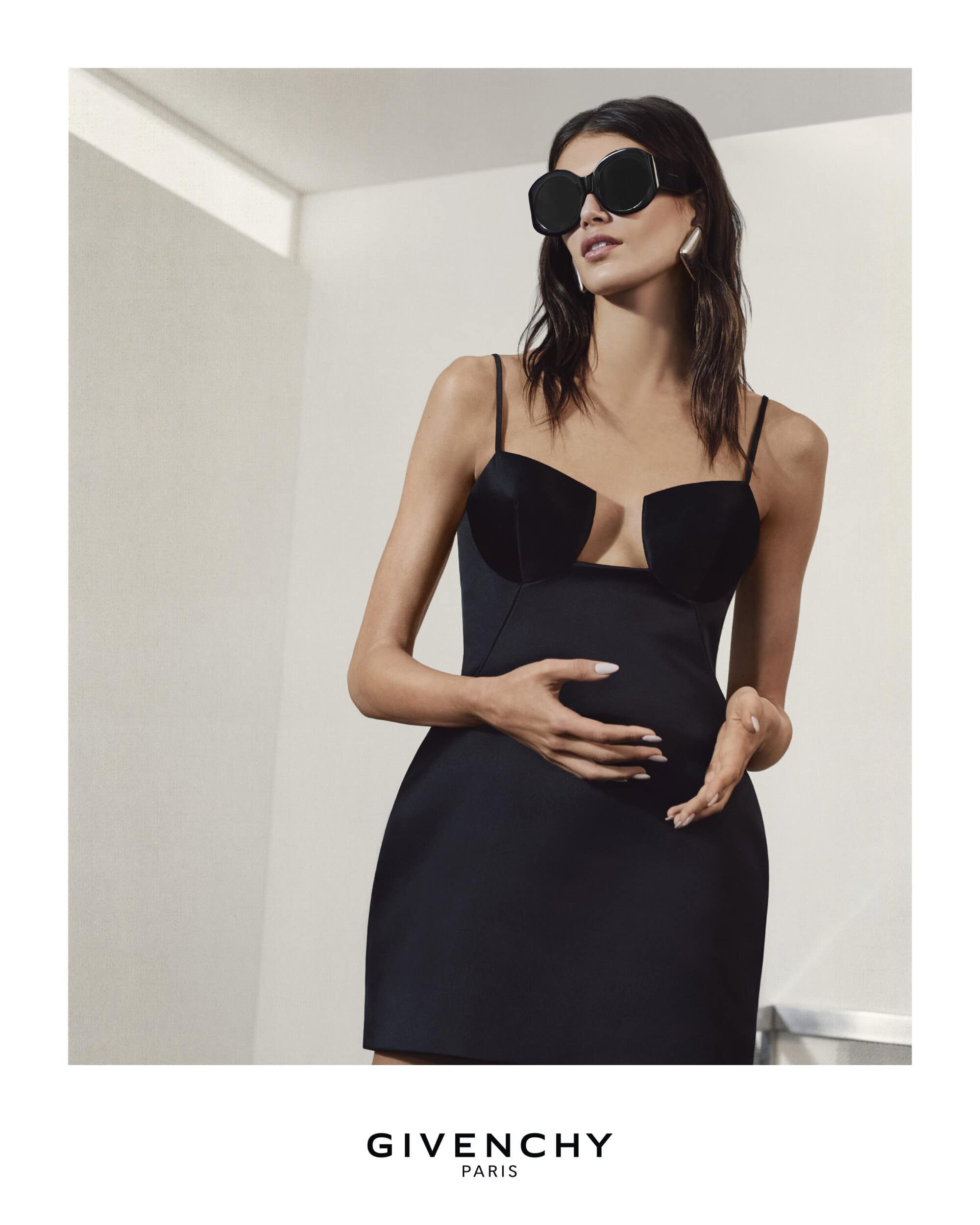 Elegant woman in a black cocktail dress and oversized sunglasses strikes a pose in an indoor setting with a modern minimalist design.