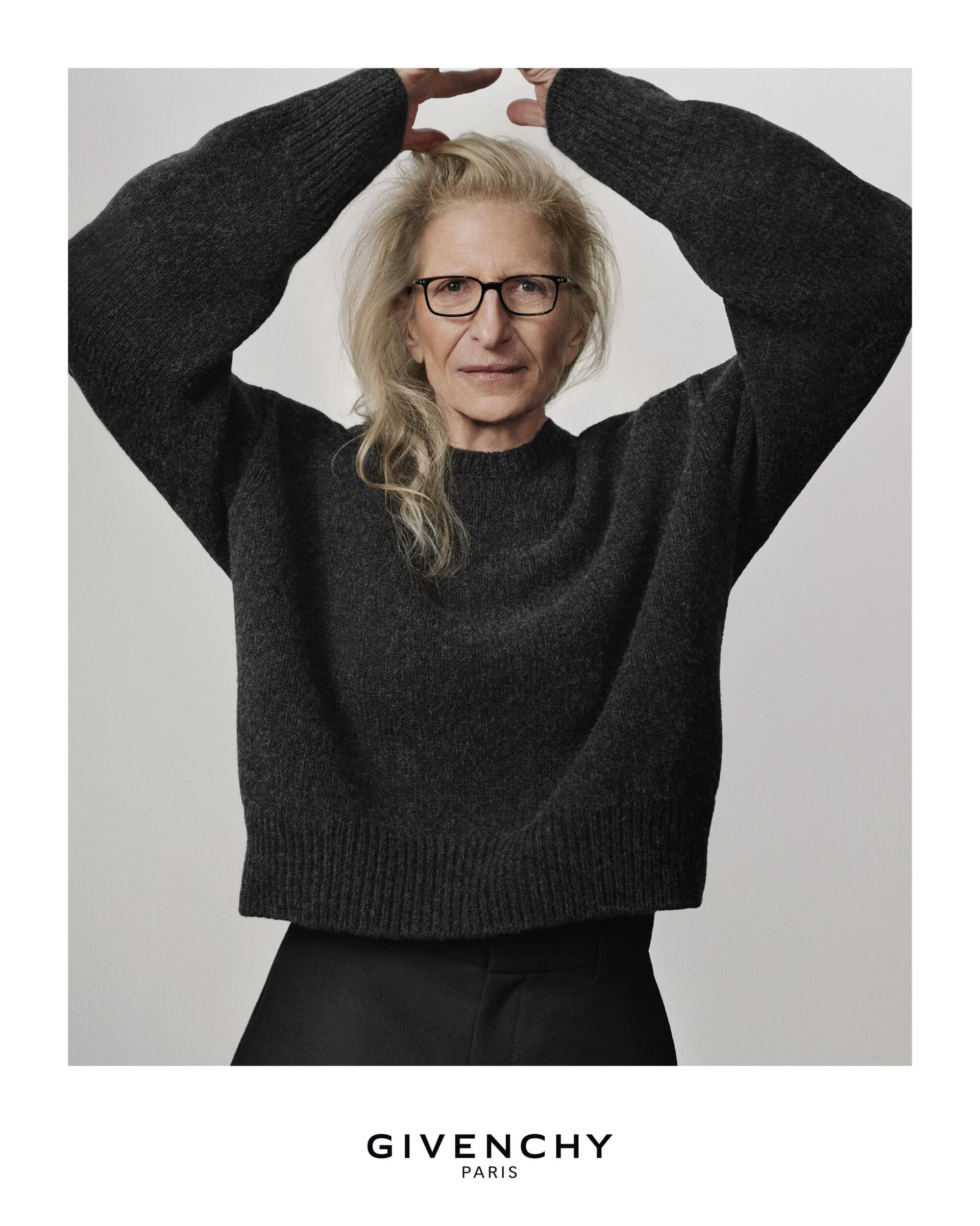 Older woman with blonde hair poses in a fashion shot, wearing a dark grey sweater against a light grey background, advertising Givenchy Paris.
