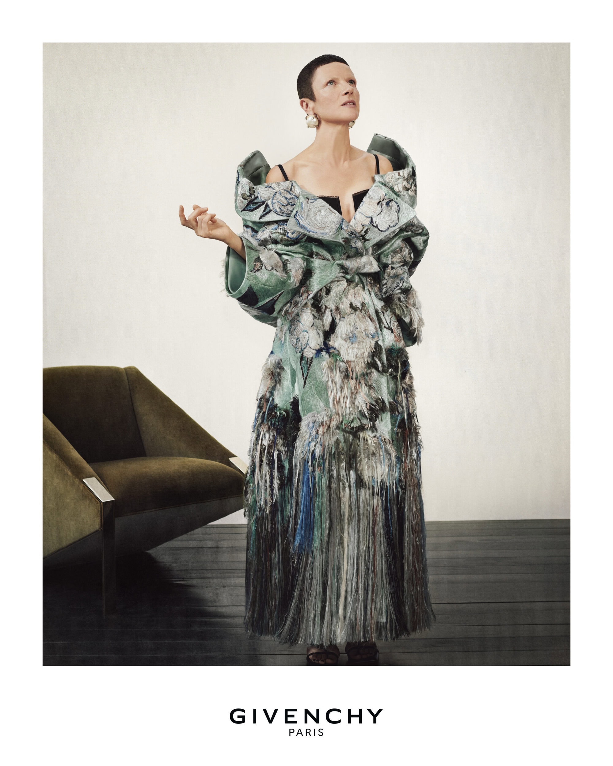 An adult female fashion model wearing a large, elaborate dress in a photo studio, posing next to a green chair for Givenchy.