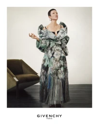 Model in a structured floral patterned gown standing against a neutral background with a green armchair.