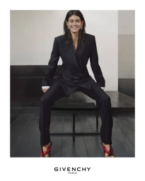 Woman in a black blazer and matching trousers seated on a low bench against a minimal studio background for Givenchy.