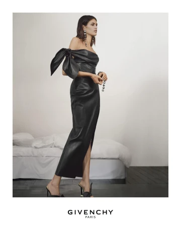 Model standing in a long black leather dress with an off-the-shoulder bow detail against a white wall.