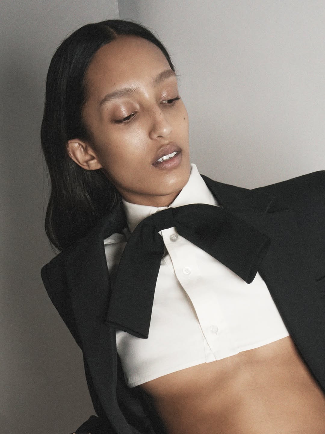 Fashion portrait of a woman with dark hair, wearing a cropped white shirt with a large black bow tie and a black blazer.