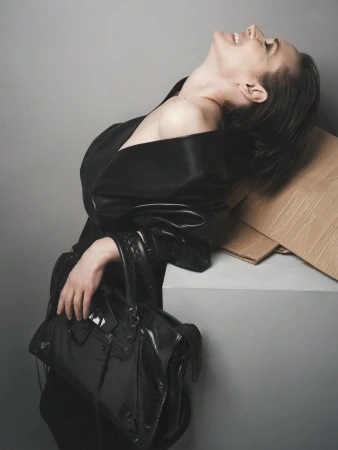 Portrait of a stylish woman laughing, wearing a black dress, holding a black bag, leaning against a gray box in a photo studio.