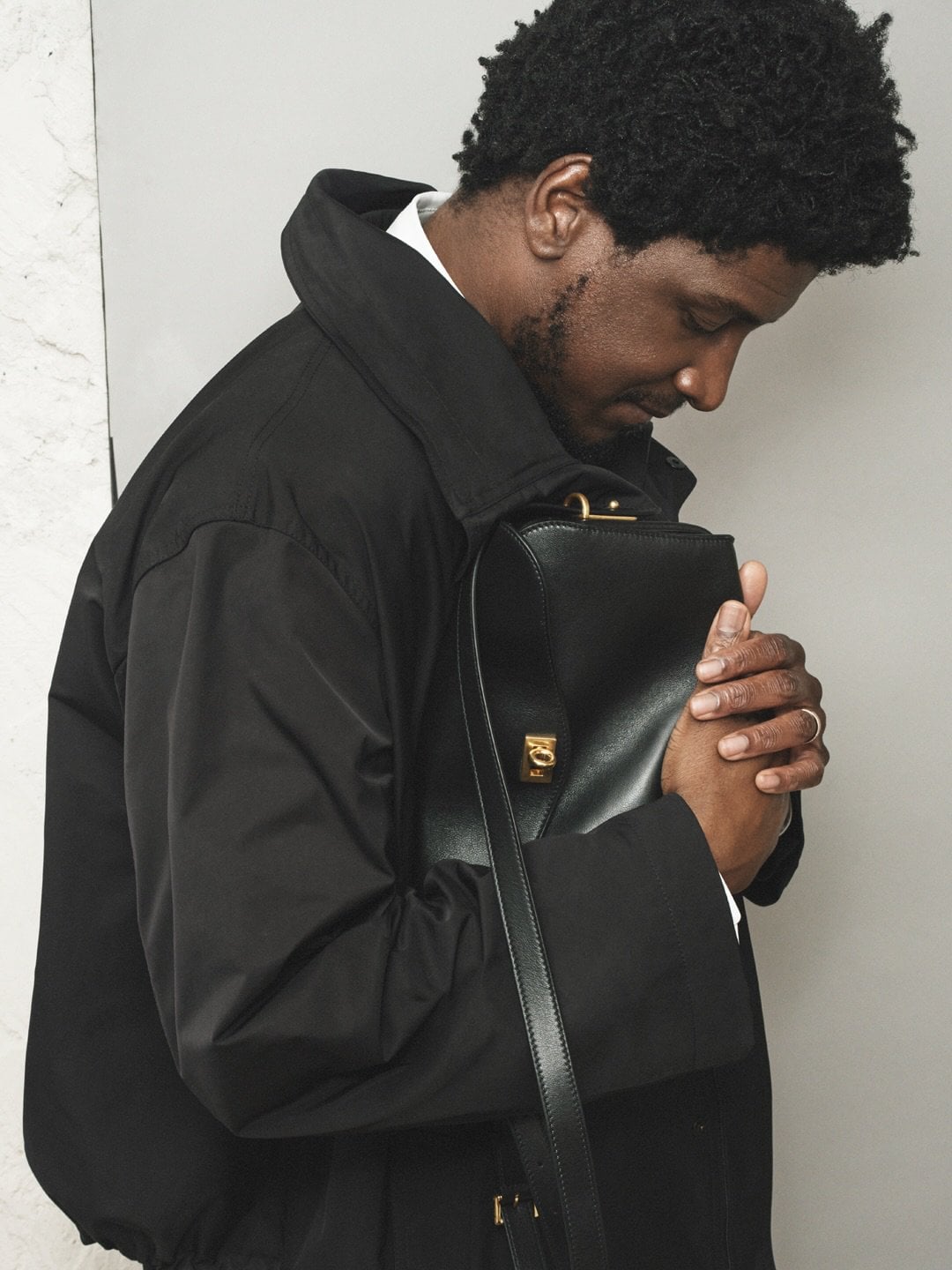 A man in a black coat holds a black leather bag against a gray studio backdrop. He wears a white shirt and has dark curly hair.