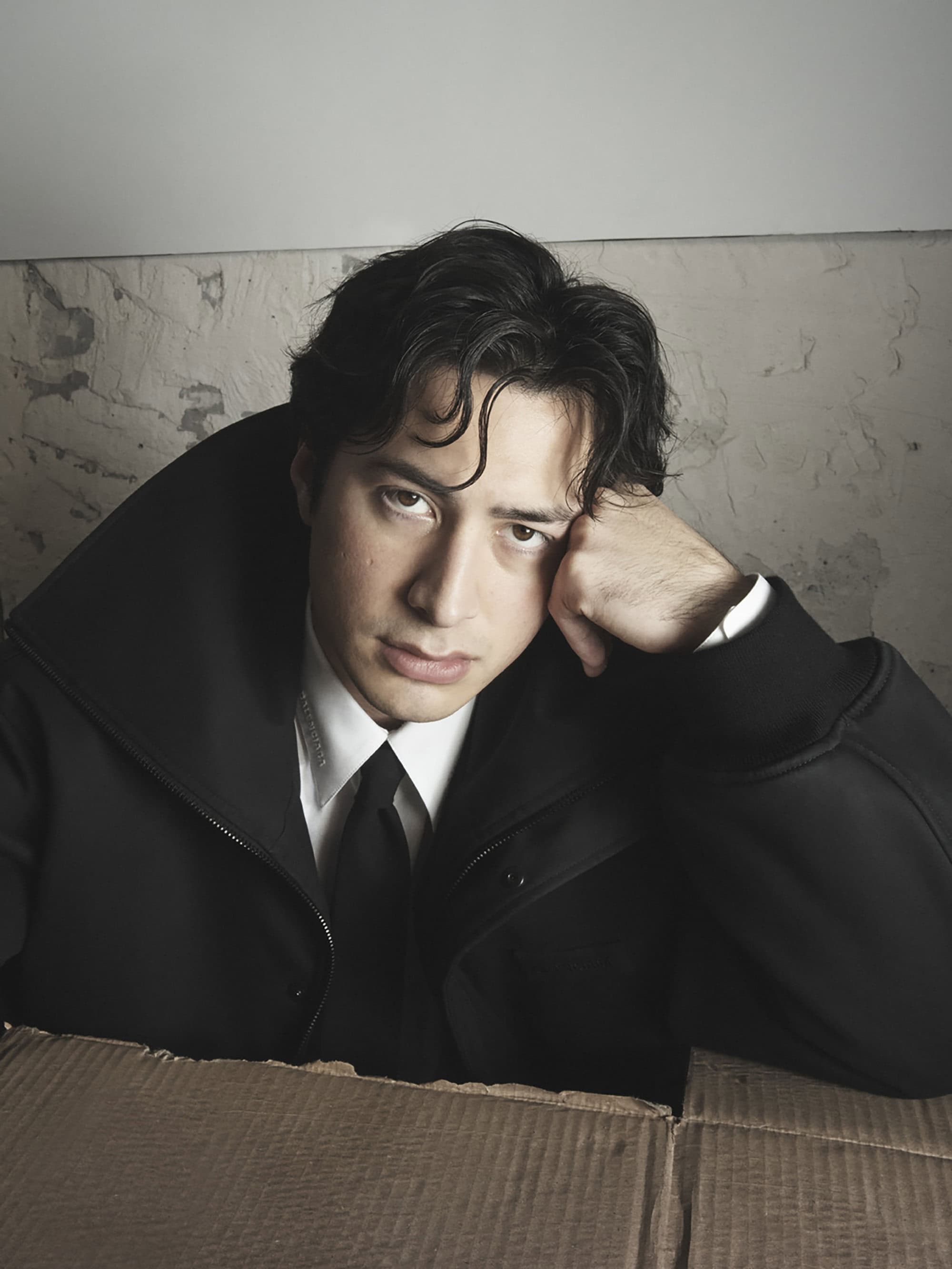Portrait of an adult male with dark hair wearing a black suit and white shirt inside of a cardboard box, posing against a wall.