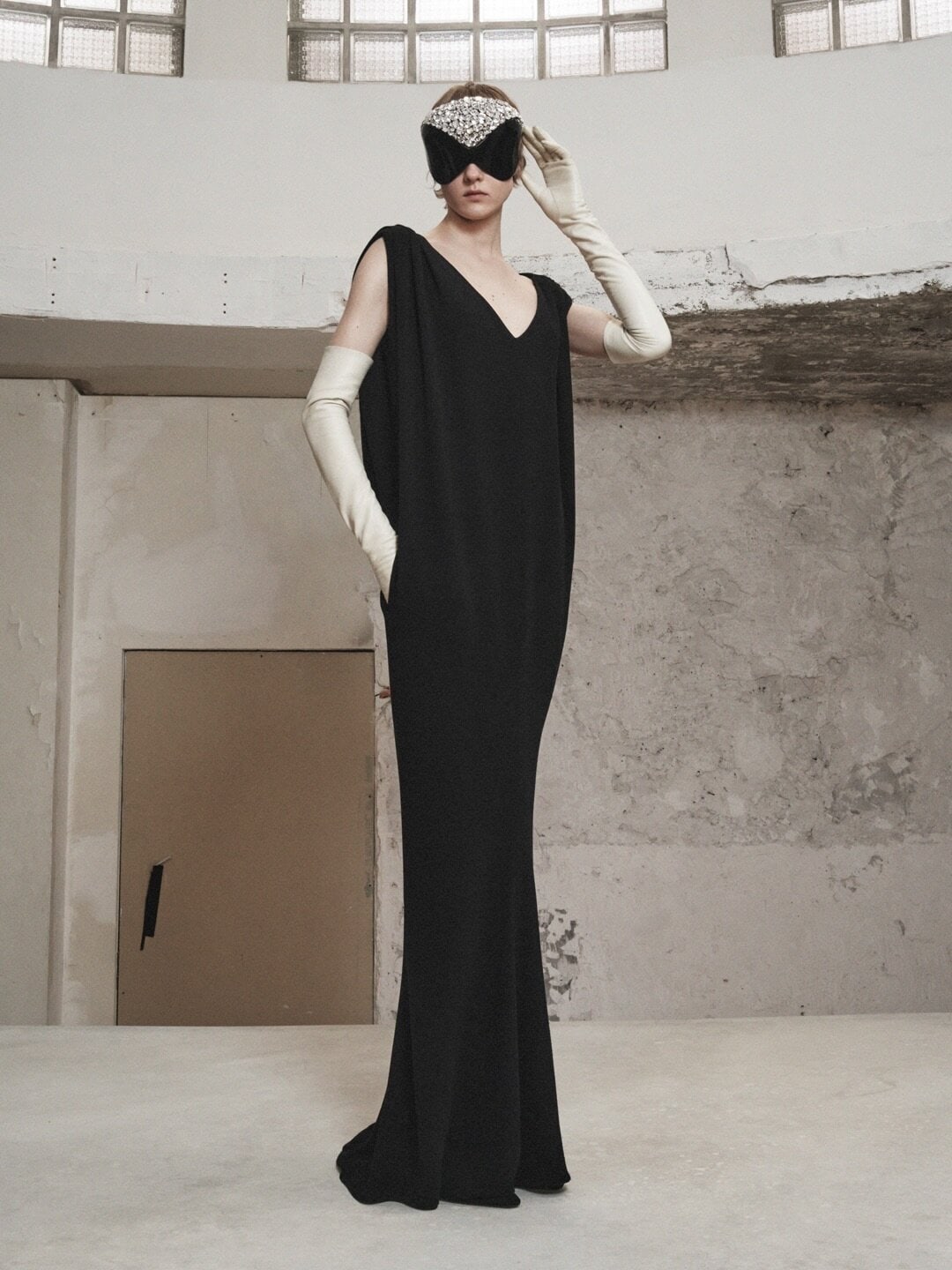 Fashion shot of a woman in a black dress and decorative mask, wearing long gloves, posing in a minimalist, industrial interior space.